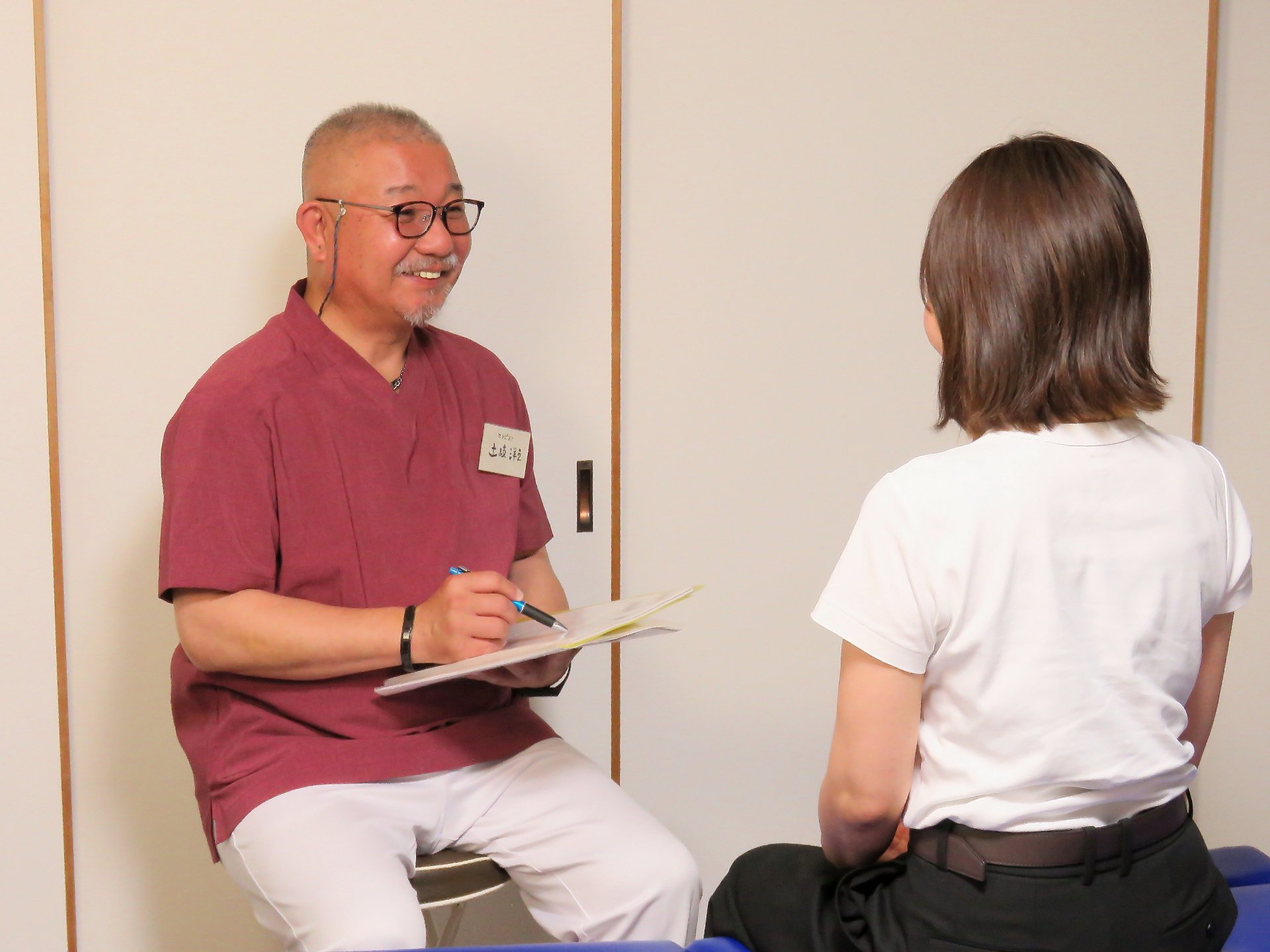 施術室でカルテを手にした院長が、椅子に座った利用者の話を落ち着いて聞いている横顔
